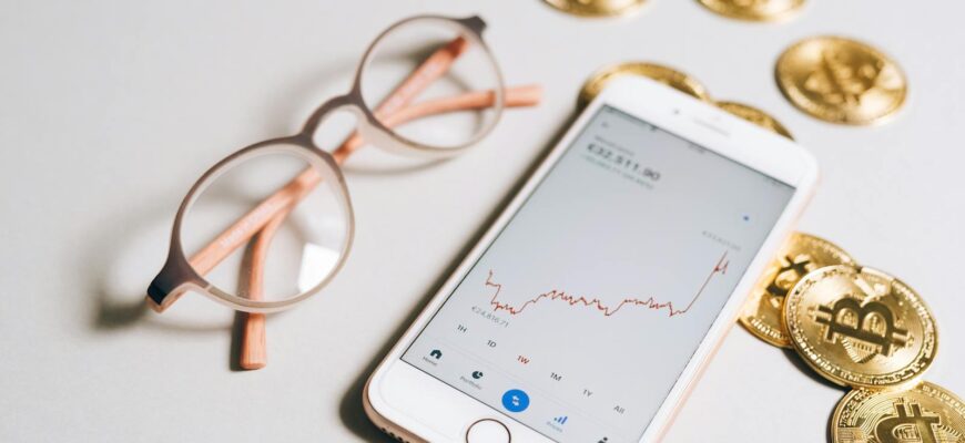 Smartphone displaying cryptocurrency graph alongside bitcoin coins and eyeglasses.